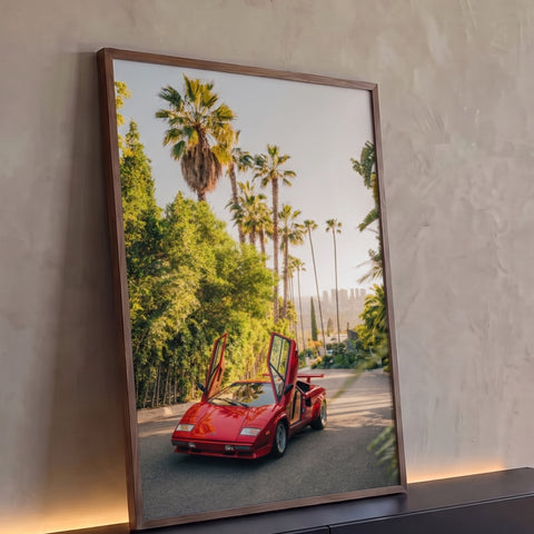 Carro esportivo vermelho anos 80 com portas abertas entre palmeiras e cidade ao fundo, quadro decorativo