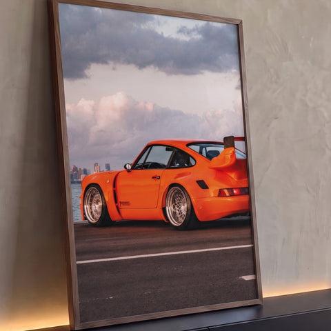 Porsche 911 laranja com aerofólio e rodas cromadas à beira-mar com skyline ao fundo, quadro decorativo