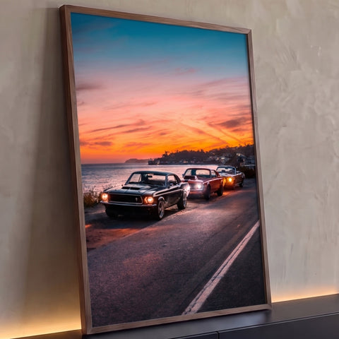 Três carros clássicos com faróis acesos em estrada à beira-mar sob céu laranja, quadro decorativo