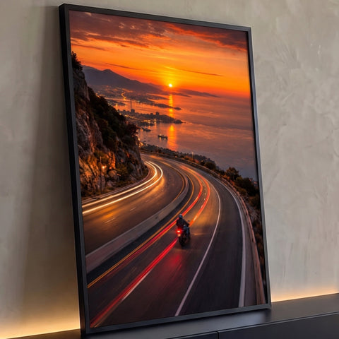 Motociclista em estrada sinuosa à beira-mar ao pôr do sol com rastros de luz e céu laranja, quadro decorativo