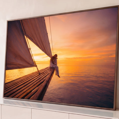 Mulher sentada na proa de veleiro com velas ao pôr do sol laranja sobre o mar calmo, quadro decorativo