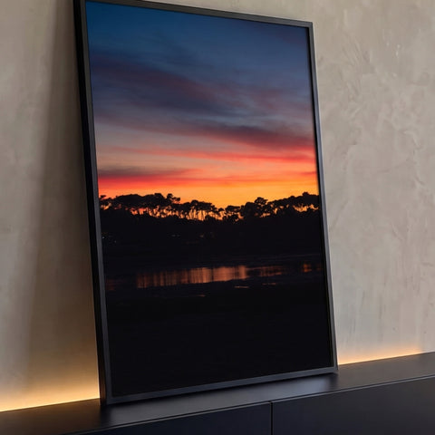 Pôr do sol laranja e azul sobre silhueta de pinheiros com reflexo na lagoa, quadro decorativo