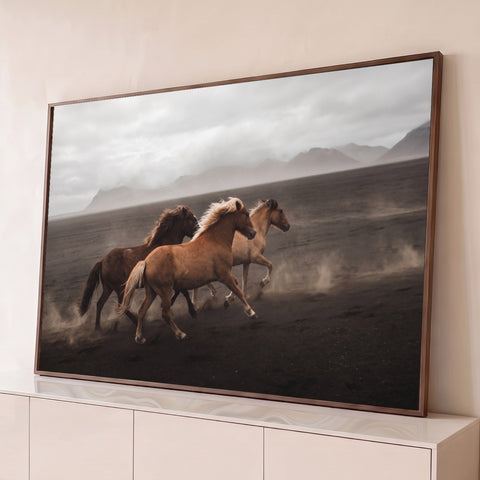 Três cavalos galopando na areia vulcânica com poeira, montanhas e céu nublado ao fundo, quadro decorativo