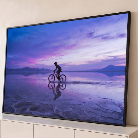 Ciclista em silhueta pedalando sobre salar com reflexo, céu roxo e rosa ao entardecer, quadro decorativo