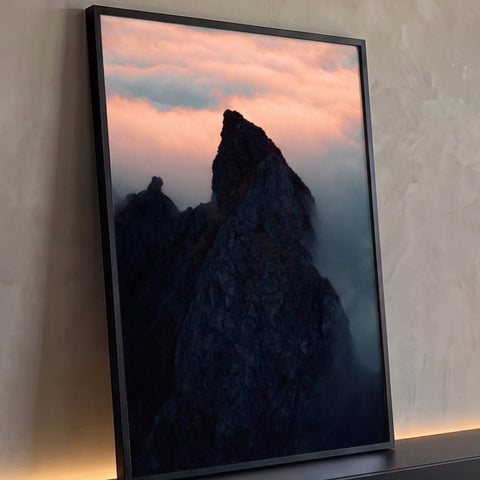 Pico de montanha escuro envolto em neblina sob céu rosa e laranja ao entardecer, quadro decorativo