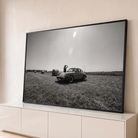 Porsche 911 clássico em campo com fardos de feno e mulher ao fundo, foto preto e branco, quadro decorativo