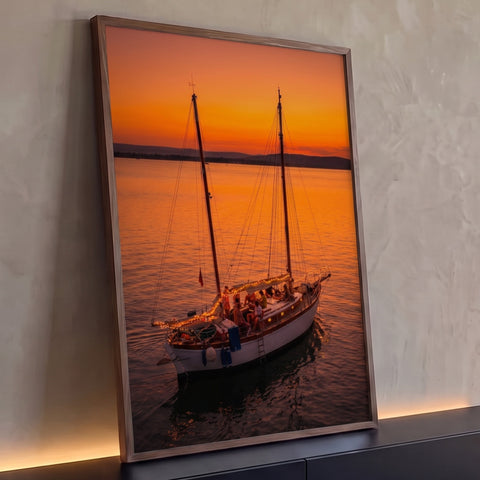 Veleiro com luzes e pessoas no convés navegando em mar calmo sob pôr do sol laranja, quadro decorativo