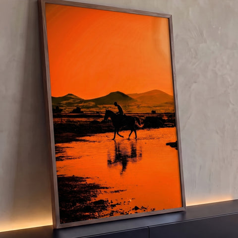 Silhueta de cavaleiro atravessando água com reflexo, montanhas e céu laranja ao pôr do sol, quadro decorativo