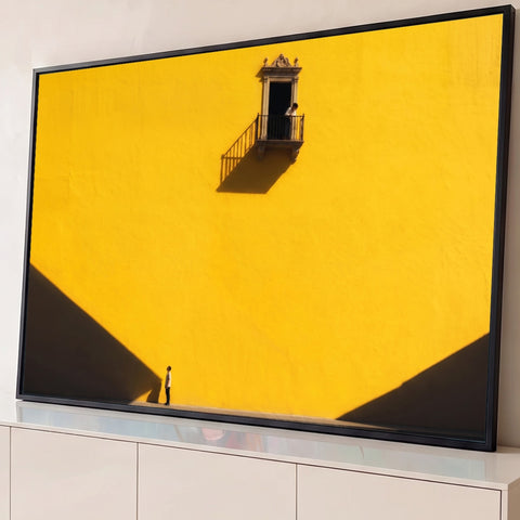 Parede amarela com sacada colonial, figura olhando de baixo e sombras diagonais, quadro decorativo