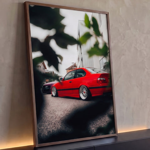 Carro coupé vermelho rebaixado com rodas prata visto entre folhas em rua urbana, quadro decorativo