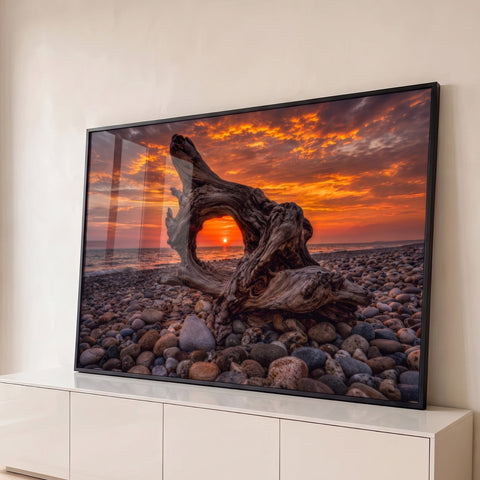 Tronco de madeira emoldurando o pôr do sol laranja sobre o mar em praia de seixos, quadro decorativo