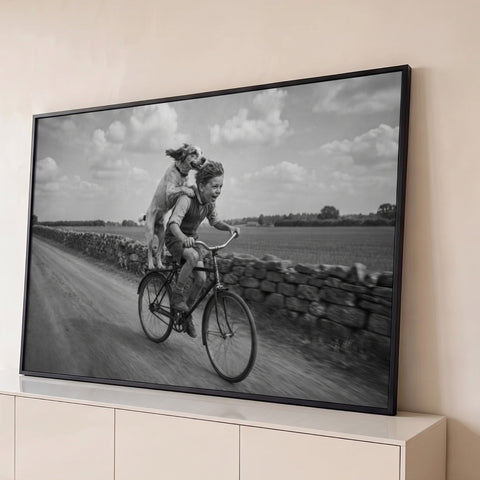 Menino sorrindo pedala bicicleta com cachorro em pé atrás, estrada rural em preto e branco, quadro decorativo