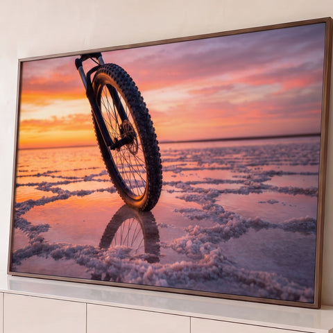 Roda de mountain bike sobre salar com reflexo na água e céu de pôr do sol laranja e rosa, quadro decorativo