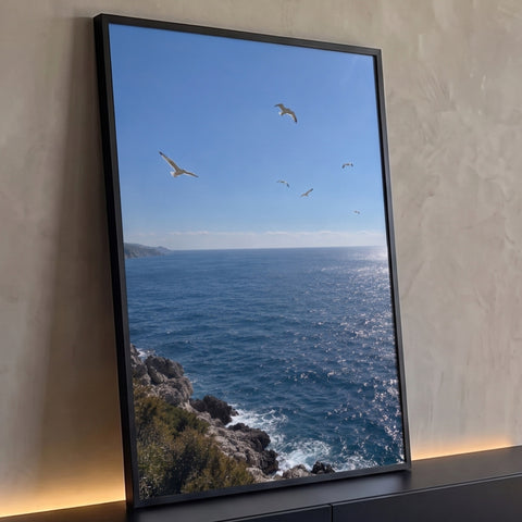 Mar azul cintilante com gaivotas voando sobre costão rochoso e vegetação, céu limpo, quadro decorativo