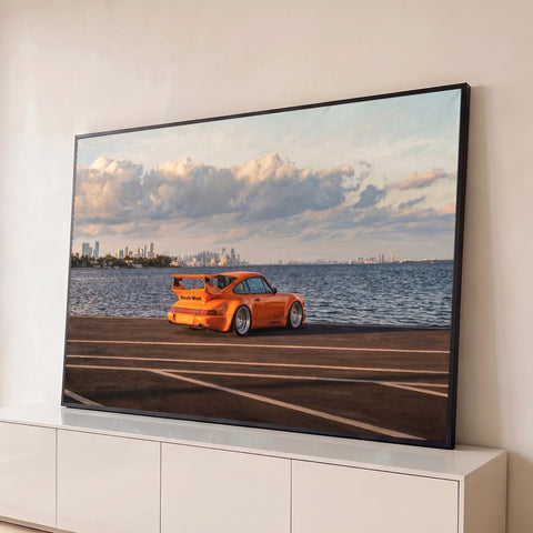 Porsche 911 laranja estacionado à beira-mar com skyline da cidade e céu com nuvens, quadro decorativo
