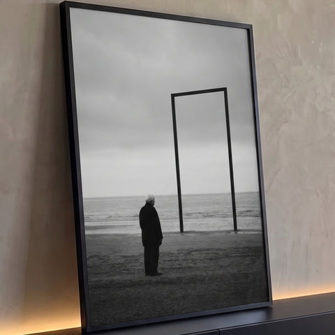 Senhor de casaco escuro olha o mar ao lado de moldura vazia na praia em preto e branco, quadro decorativo