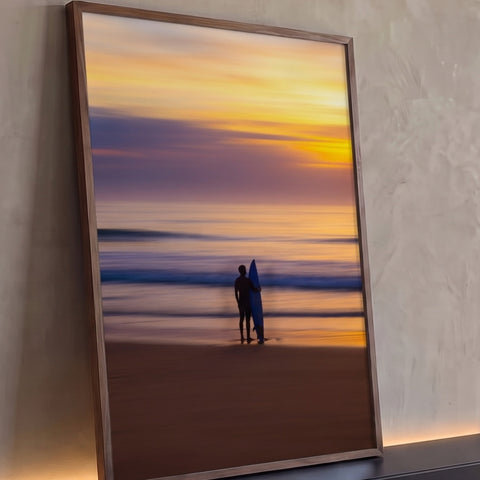 Silhueta de surfista com prancha azul na praia ao pôr do sol laranja e roxo, quadro decorativo