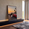Casal abraçado ao lado de Ferrari vermelha ao pôr do sol com palmeiras e mar, quadro decorativo