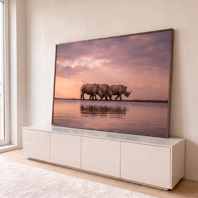 Três rinocerontes caminhando sob céu rosa ao pôr do sol, refletidos na água calma, quadro decorativo