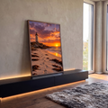 Farol branco sobre rochas, praia com pegadas e algas e pôr do sol laranja no mar, quadro decorativo