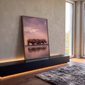 Três rinocerontes caminhando à beira do lago com reflexo e céu rosa ao pôr do sol, quadro decorativo