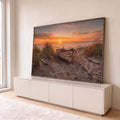 Barco de madeira rústico na areia entre capim de dunas com pôr do sol laranja no mar, quadro decorativo