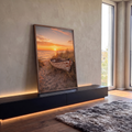 Barco de madeira descascado entre dunas e capim na praia, sob pôr do sol laranja no mar, quadro decorativo