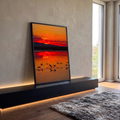 Flamingos em silhueta voando sobre lago refletindo pôr do sol laranja e vermelho, quadro decorativo