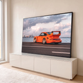 Porsche 911 laranja com aerofólio à beira-mar, prédios da cidade e nuvens rosadas ao fundo, quadro decorativo