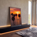 Pôr do sol laranja no mar com palmeiras altas em silhueta e veleiro branco ao centro, quadro decorativo