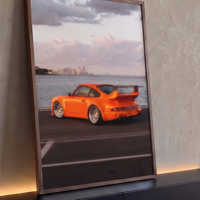 Porsche 911 laranja com aerofólio e rodas raiadas à beira-mar, skyline ao fundo, quadro decorativo