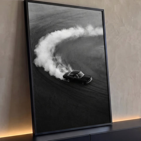 Carro clássico preto derrapando na areia com rastro de fumaça branca, vista aérea em P&B, quadro decorativo