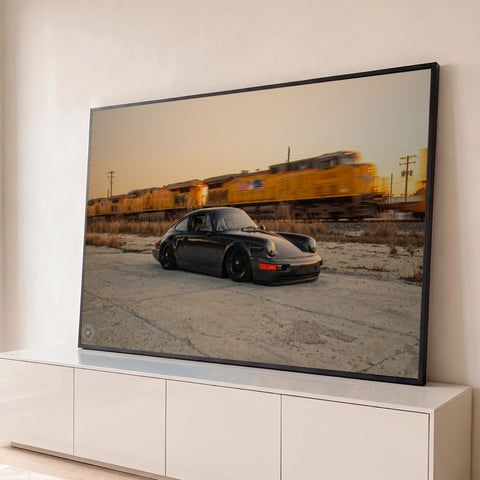 Porsche 911 preto rebaixado diante de trem de carga amarelo borrado sob céu de entardecer, quadro decorativo