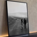 Casal de mãos dadas caminhando em estrada de deserto com carro ao fundo em preto e branco, quadro decorativo