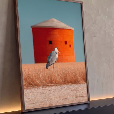 Garça-cinzenta em campo de grama dourada com silo laranja e céu azul ao fundo, quadro decorativo
