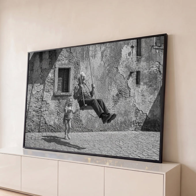 Avô sorridente num balanço e neta brincando em rua de pedra, fotografia preto e branco, quadro decorativo