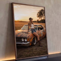 Mulher de boné e botas apoiada em muscle car enferrujado ao pôr do sol no campo, quadro decorativo