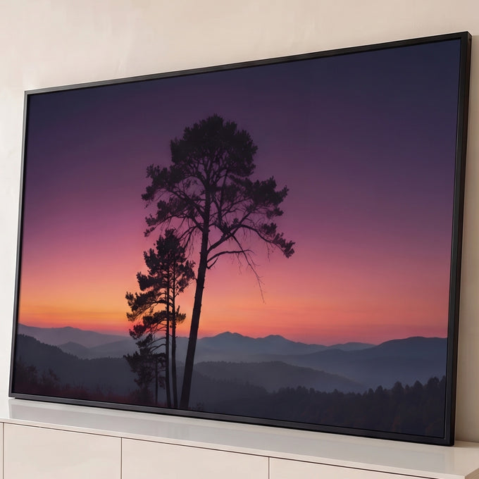 Pinheiros em silhueta sob céu roxo e laranja ao pôr do sol e montanhas enevoadas, quadro decorativo