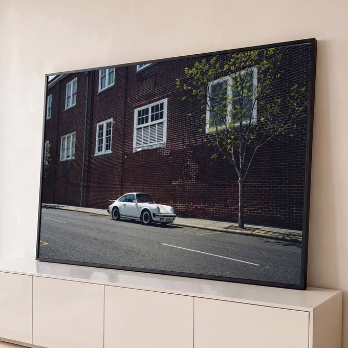 Porsche 911 clássico branco estacionado em rua diante de prédio de tijolos vermelhos, quadro decorativo
