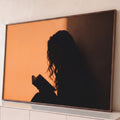 Silhueta de mulher de cabelos cacheados lendo livro em contraluz sobre parede laranja, quadro decorativo