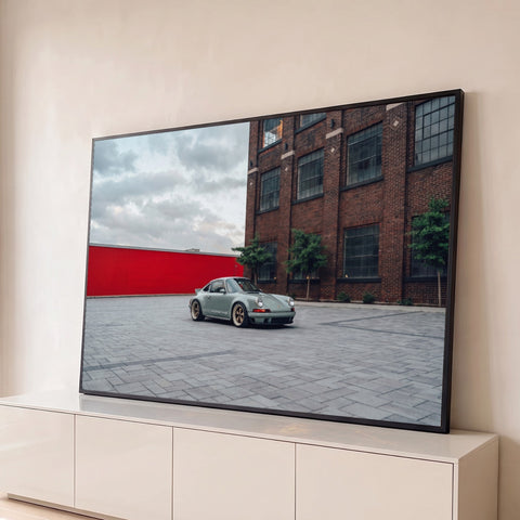Porsche 911 verde sage em pátio de pedra diante de prédio de tijolos e muro vermelho, quadro decorativo