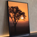 Árvore em silhueta com sol entre os galhos em céu laranja sobre colinas enevoadas, quadro decorativo