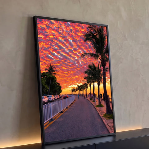 Pôr do sol laranja e vermelho sobre orla com palmeiras, ciclistas e mar ao fundo, quadro decorativo