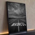 Estrada com a palavra MADNESS pintada no asfalto sob céu tempestuoso em preto e branco, quadro decorativo
