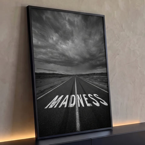 Estrada com a palavra MADNESS pintada no asfalto sob céu tempestuoso em preto e branco, quadro decorativo