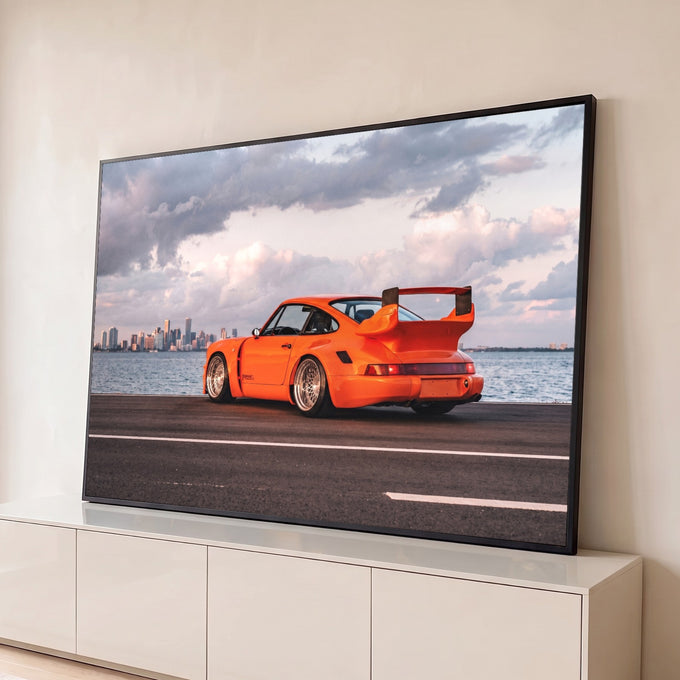 Porsche 911 laranja com aerofólio à beira-mar, prédios da cidade e nuvens rosadas ao fundo, quadro decorativo