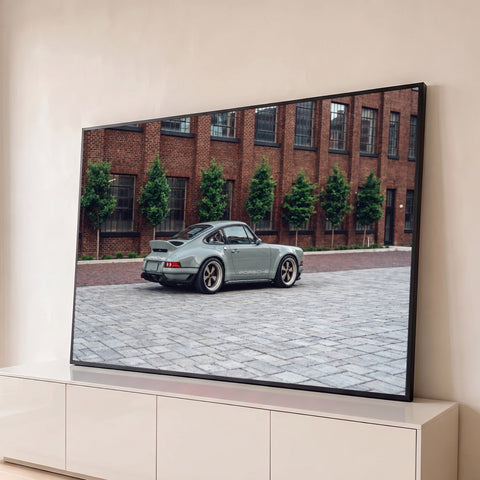 Porsche 911 clássico cinza com rodas douradas em rua de paralelepípedos e prédio de tijolos, quadro decorativo