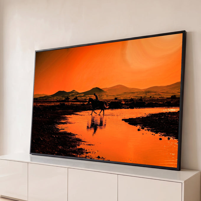 Silhueta de cavaleiro cruzando rio com reflexo na água, montanhas e céu laranja ao pôr do sol, quadro decorativo