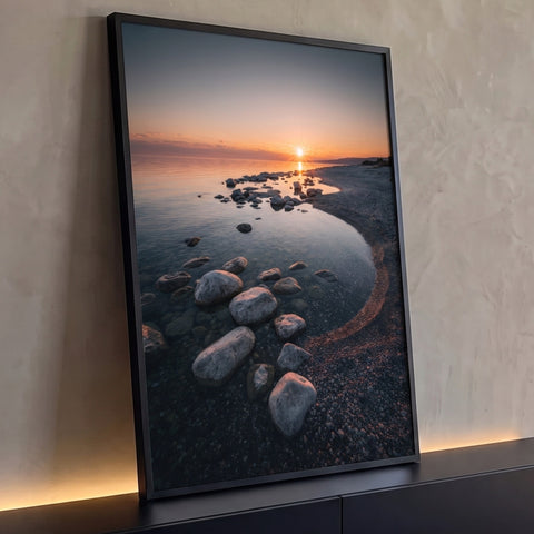 Pôr do sol laranja sobre água calma com pedras e cascalho na margem, quadro decorativo