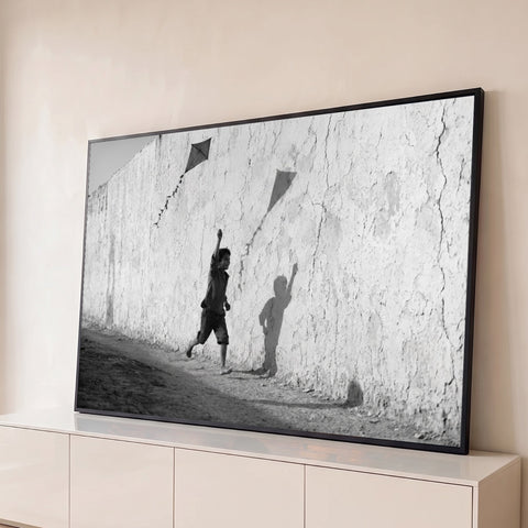 Menino descalço correndo com pipa junto a muro rachado, sombra na parede, foto P&B, quadro decorativo